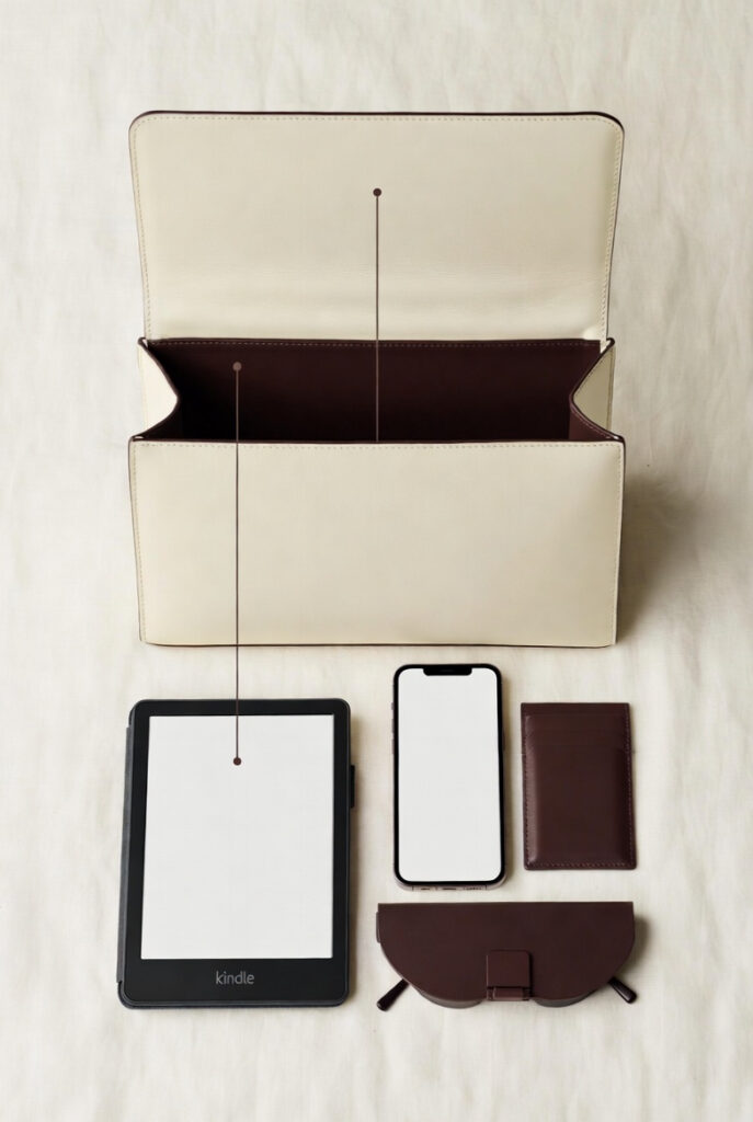 Technical flat-lay of a Kindle, smartphone, and essentials fitting horizontally inside a Luruge East-West handbag.